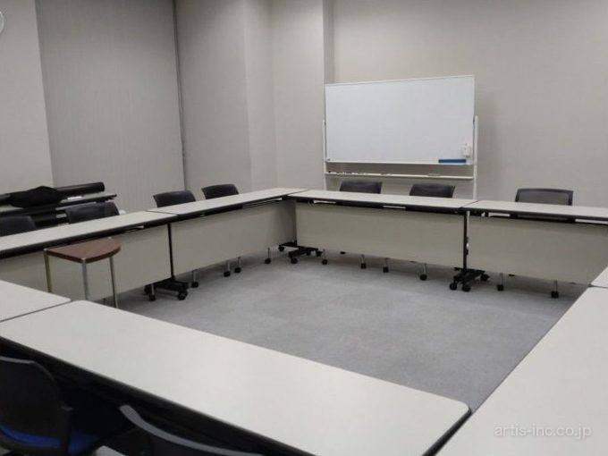 灰色の壁の会議室。白い長机がコの字に並び、奥にホワイトボード